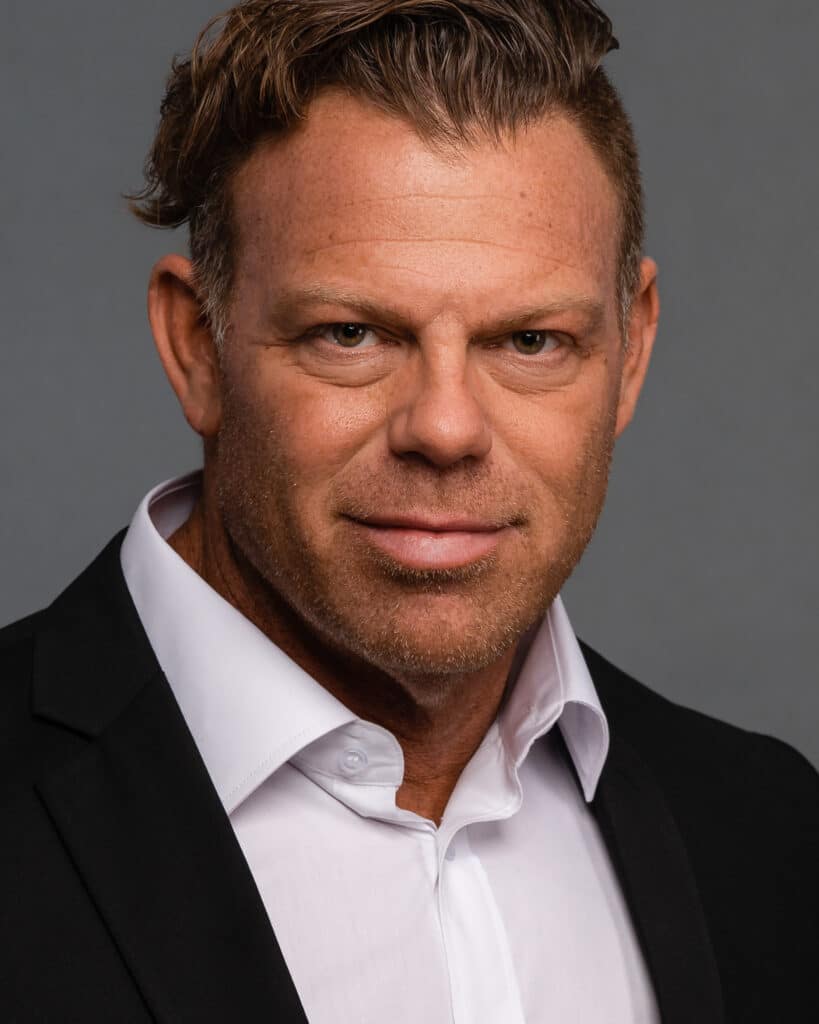 Mike Fortin, known for his roles in Ozark, Team Seal, and Jack Ryan, poses for a professional headshot. He has a confident expression, with short, neatly styled hair and a well-groomed beard. He wears a dark blazer over a light shirt, standing against a neutral background that highlights his sharp features and intense gaze.