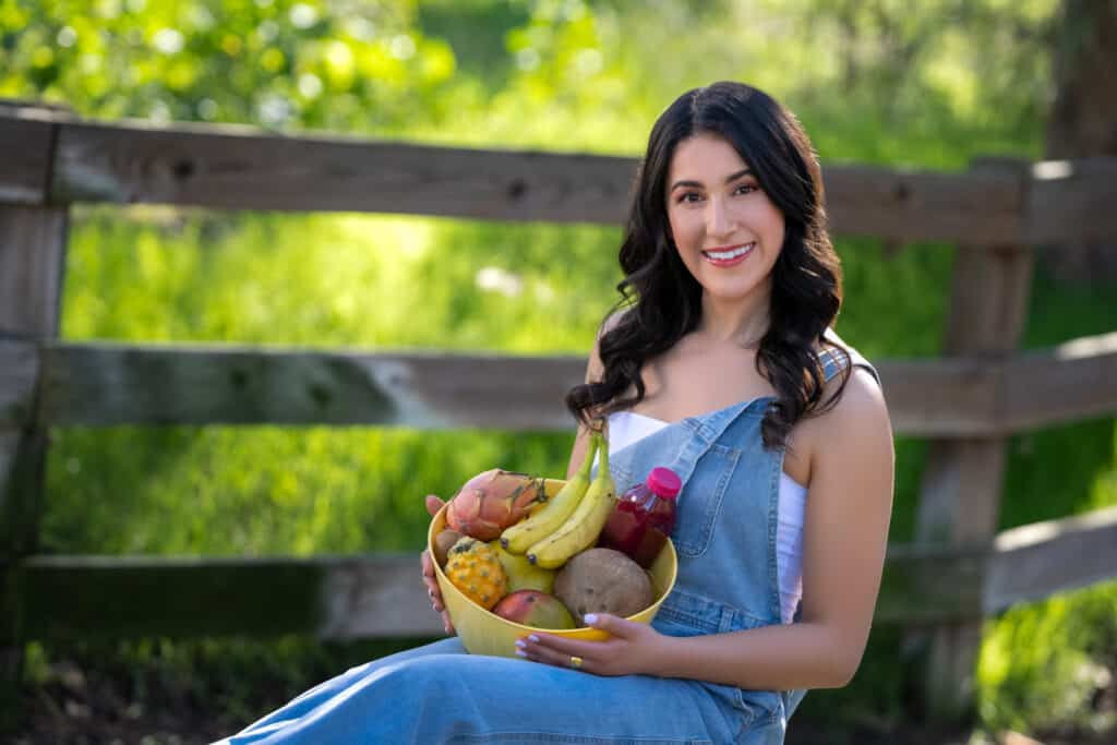 A healthy lifestyle portrait of a woman holding a basket of fresh fruits. She is dressed in casual, comfortable clothing, standing outdoors in a sunny garden. The woman smiles warmly, radiating health and happiness. The basket is filled with a colorful assortment of fruits, including apples, oranges, and bananas, symbolizing her commitment to a nutritious diet.