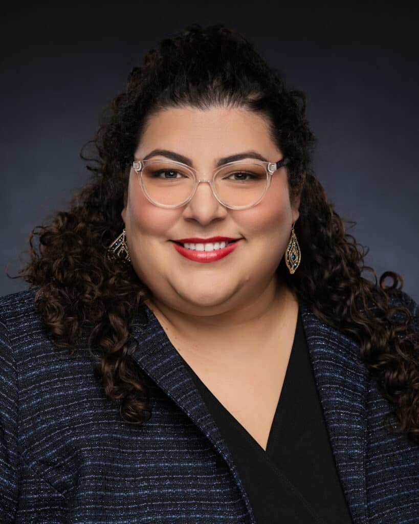 Rebecca, a professional educator, smiles warmly at the camera. She is dressed in a smart, professional outfit, exuding confidence and approachability. Her background features a bookshelf filled with educational materials, emphasizing her dedication to teaching and learning