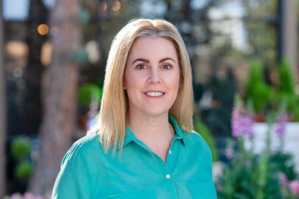 A professional headshot of Carrie, taken outdoors in natural light for the CDM Home Tour in Newport Beach, CA. Carrie is dressed in elegant attire, smiling warmly, with a scenic background of greenery and sunlight, exuding confidence and approachability.